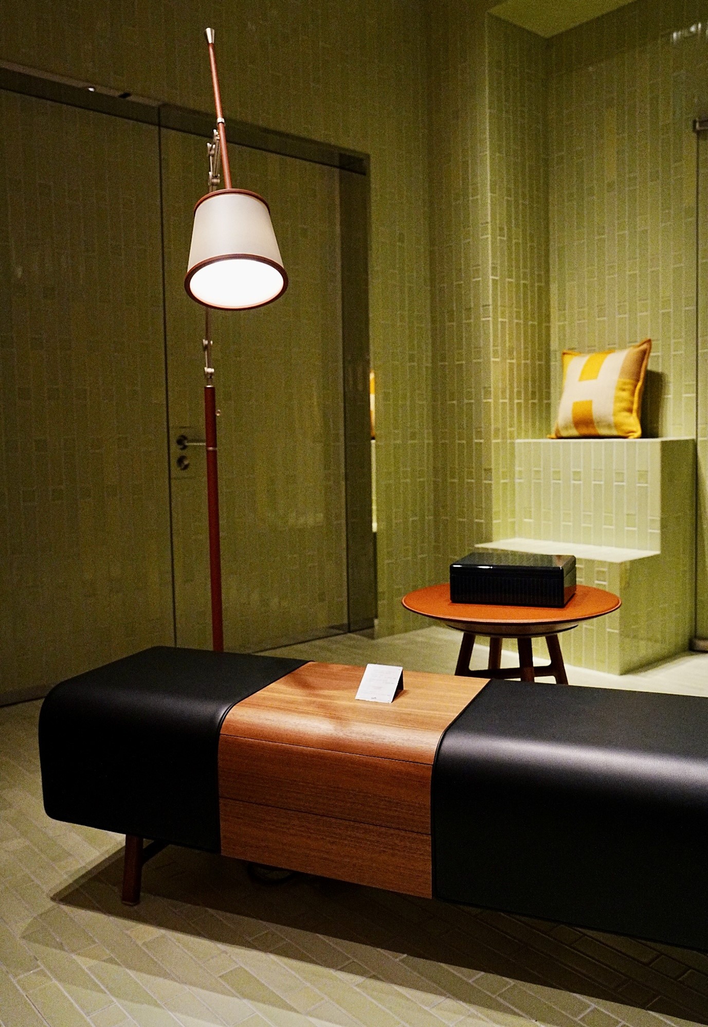 Modern hotel lobby with a black leather and timber bench, tall floor lamp, and small side table holding a box against an olive green textured wall