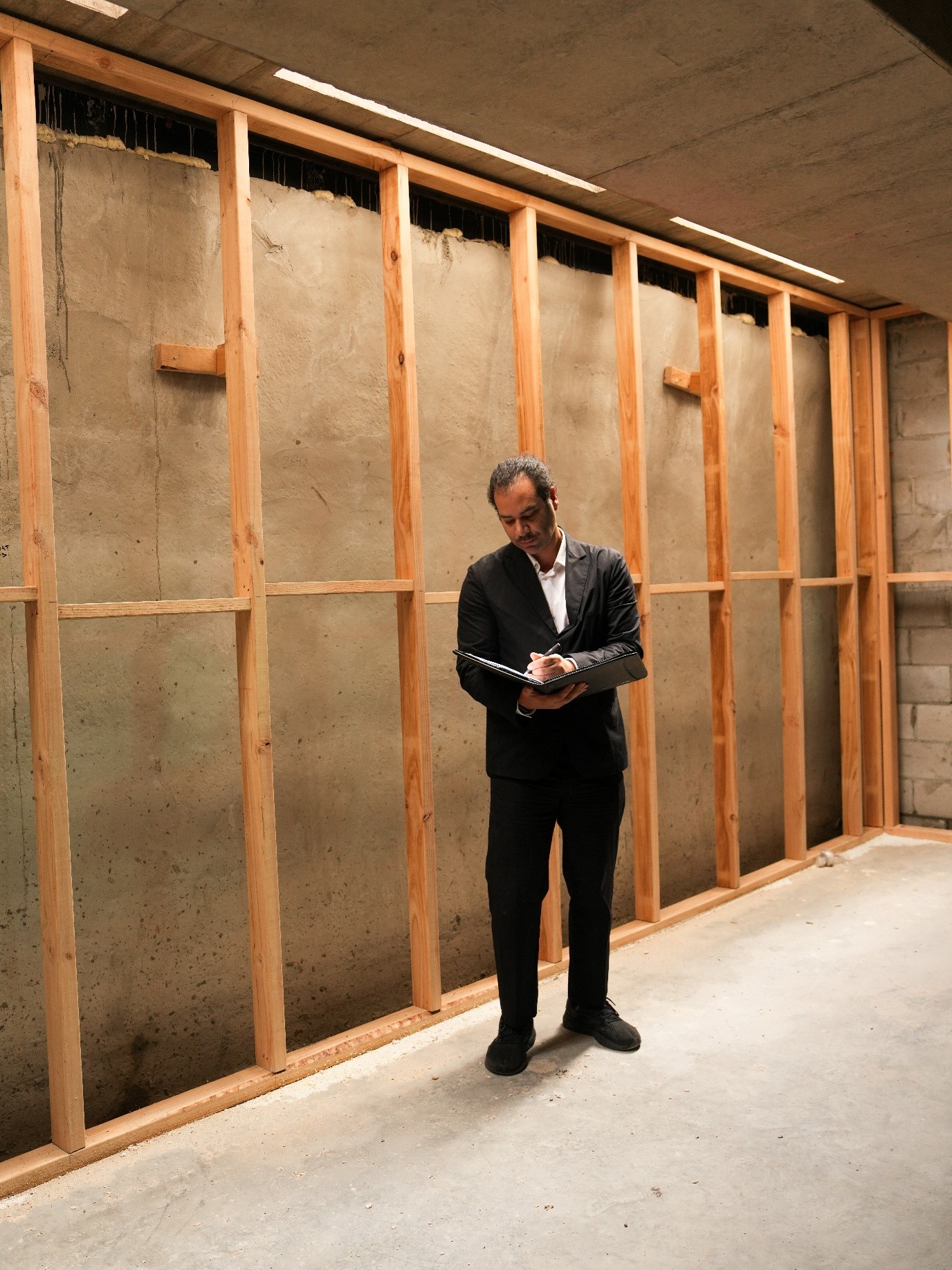 Building inspector in a black suit writing notes on a clipboard inside a property with exposed timber wall framing and concrete floor