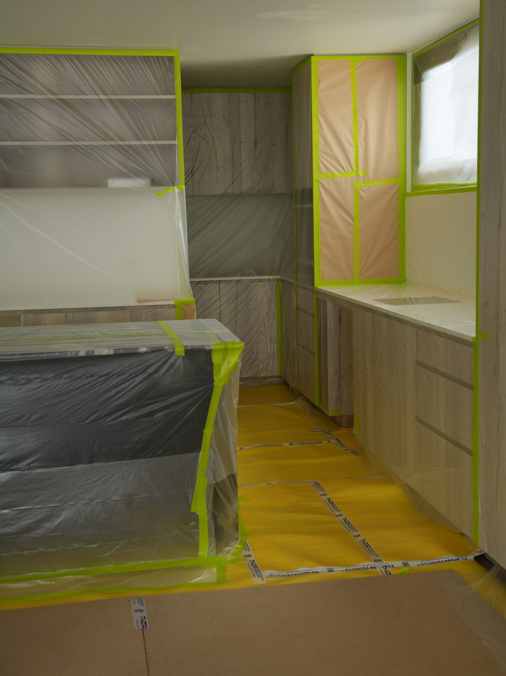 Kitchen prepped for spray painting, with cabinetry, benchtops, and floors covered in plastic sheeting and green masking tape