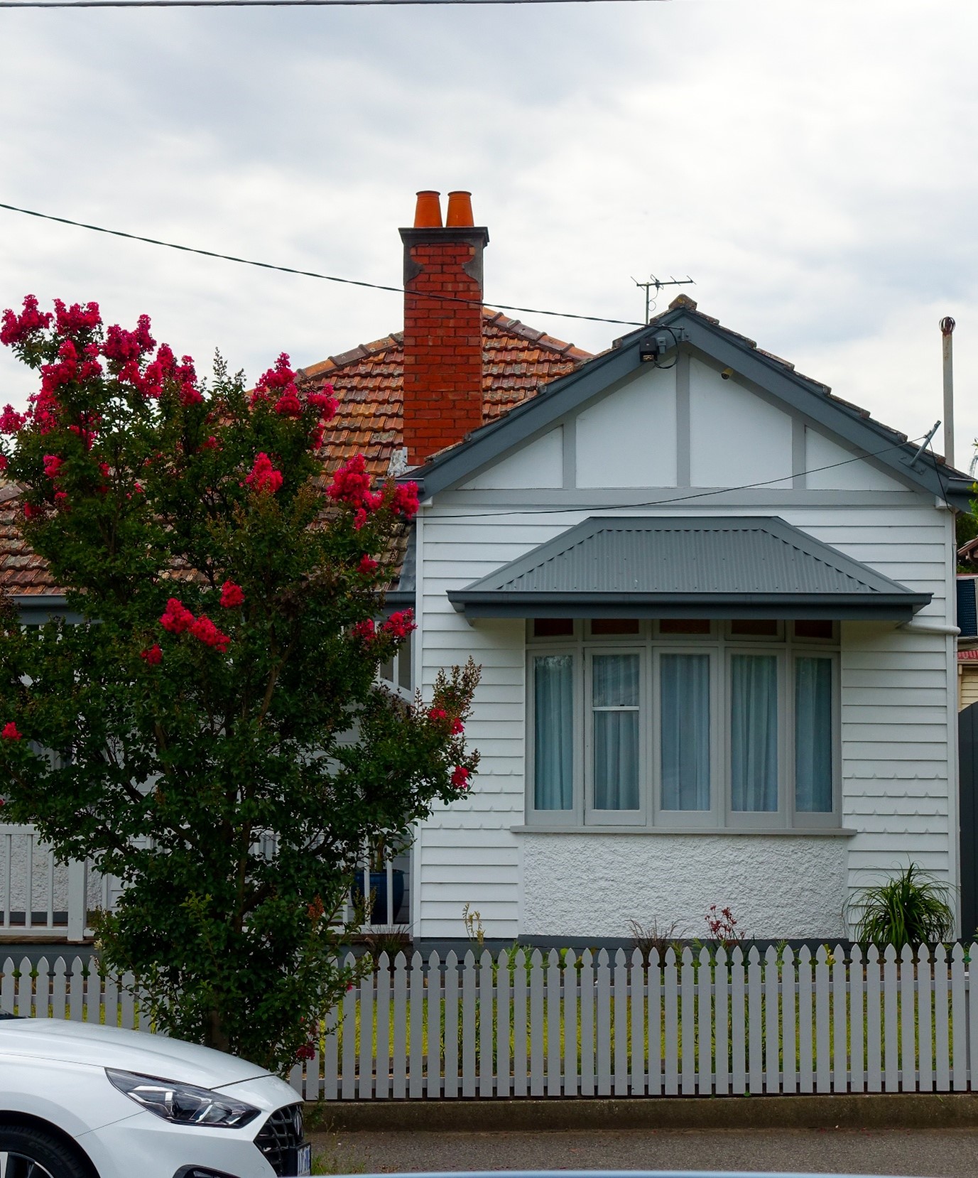 White weatherboard home with a grey tiled roof, red brick chimney, bay window, and a flowering crepe myrtle tree behind a white picket fence